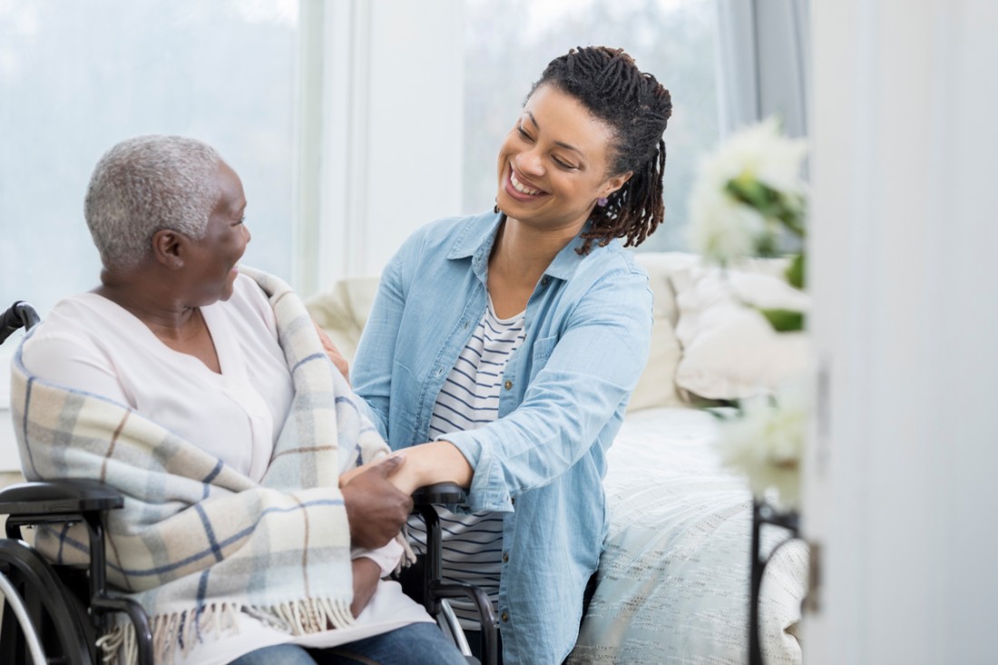 A smiling female care giver holds the hand of a senior woman who is in a wheelchair at home