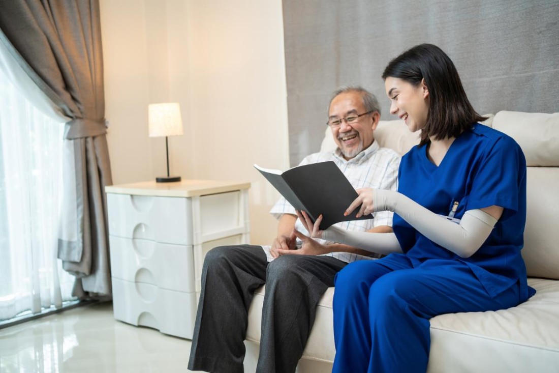 Caregiver sitting with an elderly man reading a book