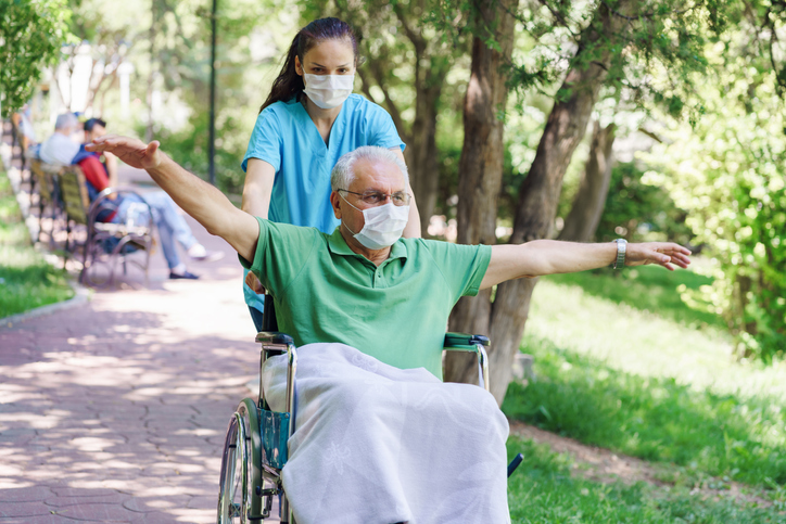 Young female caregiver pushing happy older man in wheelchair through the park wearing masks.