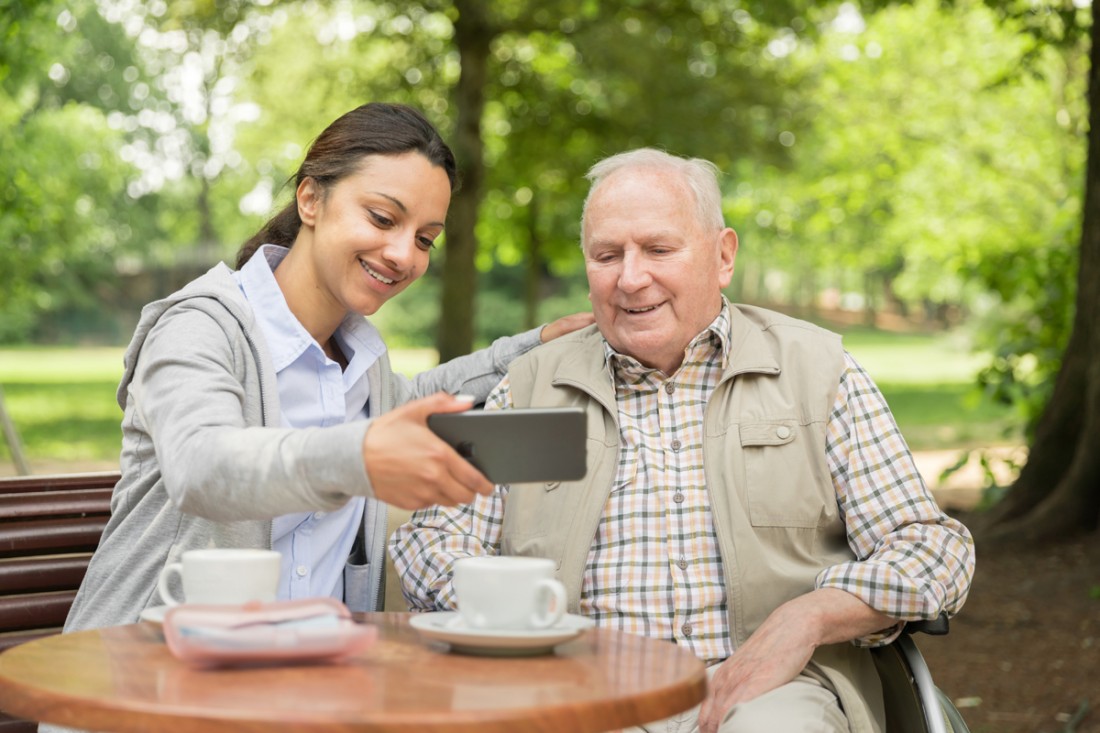 A young female caregiver and a senior man sitting in his wheelchair are having coffee in the park and taking a selfie