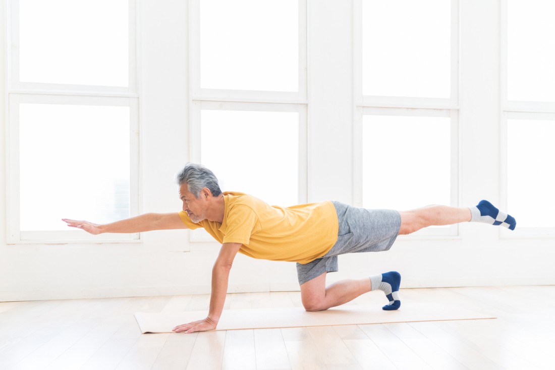 An older man does a bird-dog or opposite leg and arm raise on a yoga mat