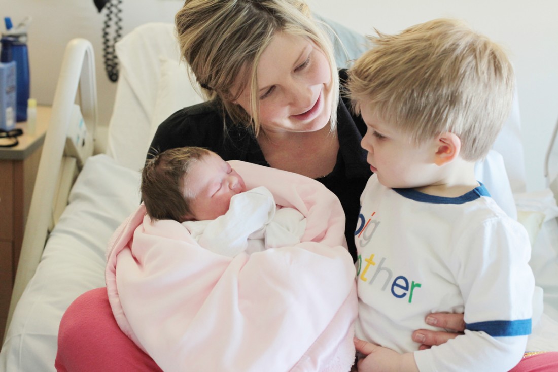 A smiling mom sits in her hospital bed holding her toddler son and newborn baby