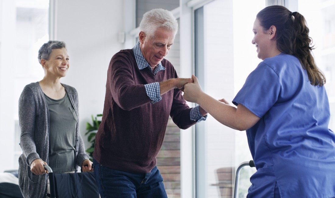 A smiling female caregiver assists a senior man to stand from his wheelchair while a happy female looks on