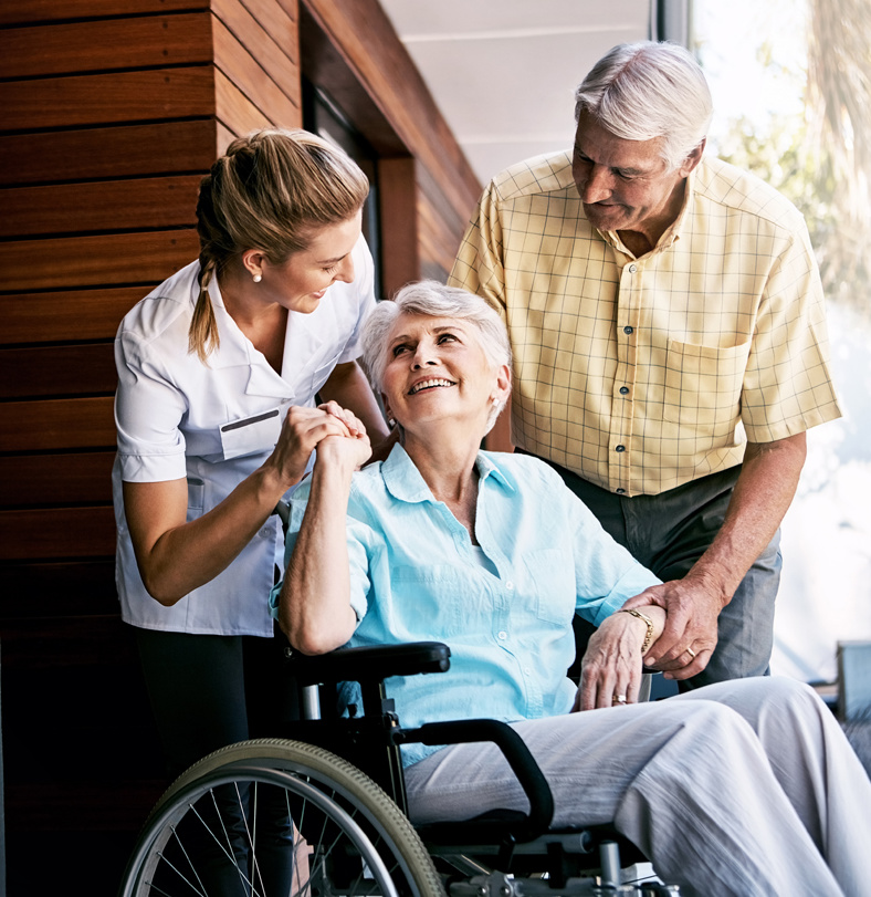 A respite care worker and a senior male hold the hands of a smiling senior woman in a wheelchair