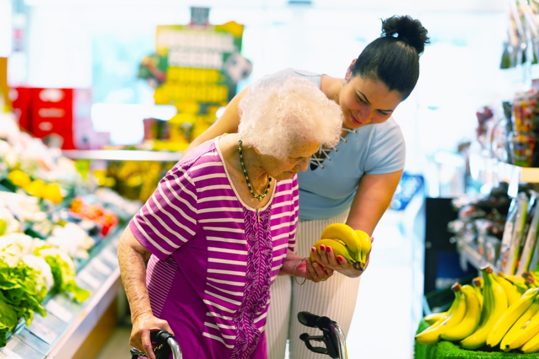  A smiling female caregiver supports a senior woman with a walker to select bananas while grocery shopping