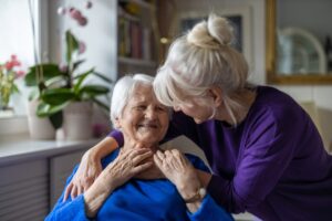 An older lady puts her arm around an older lady who is sitting down and talking to