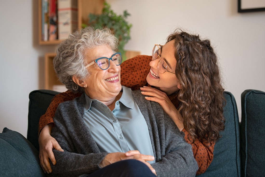 A younger lady laughs with an older woman sitting on a couch.