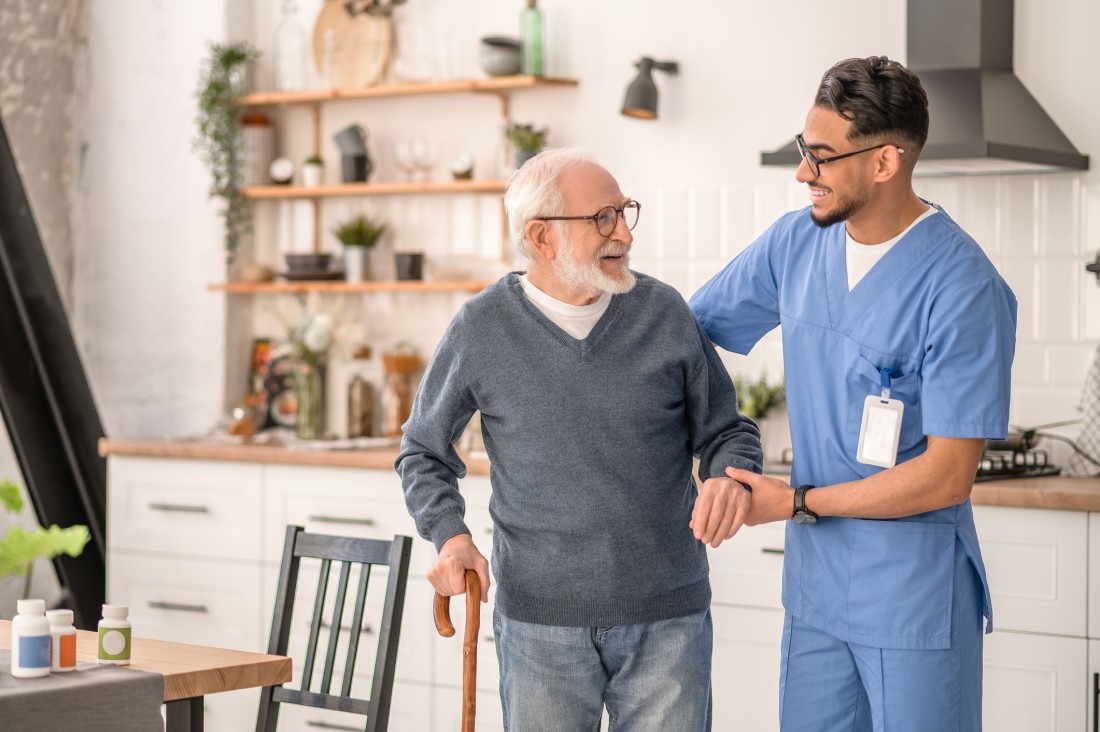 A male nurse helps an older gentleman walking with a cane through a home kitchen.