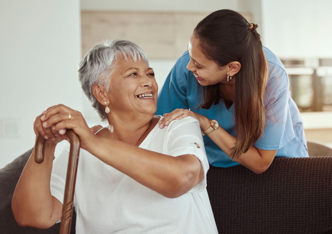 A nurse leans over the couch and happily talks to the older lady sitting there with her cane.