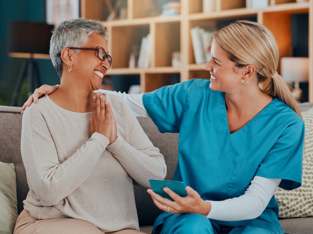 A female nurse talks happily with an older lady while sitting in a living room.